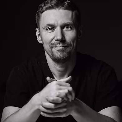 A professional portrait of Ihor Kachula LMT. He is wearing a dark scrub top, with his hands clasped in front of him and is smiling into the camera against a dark background.